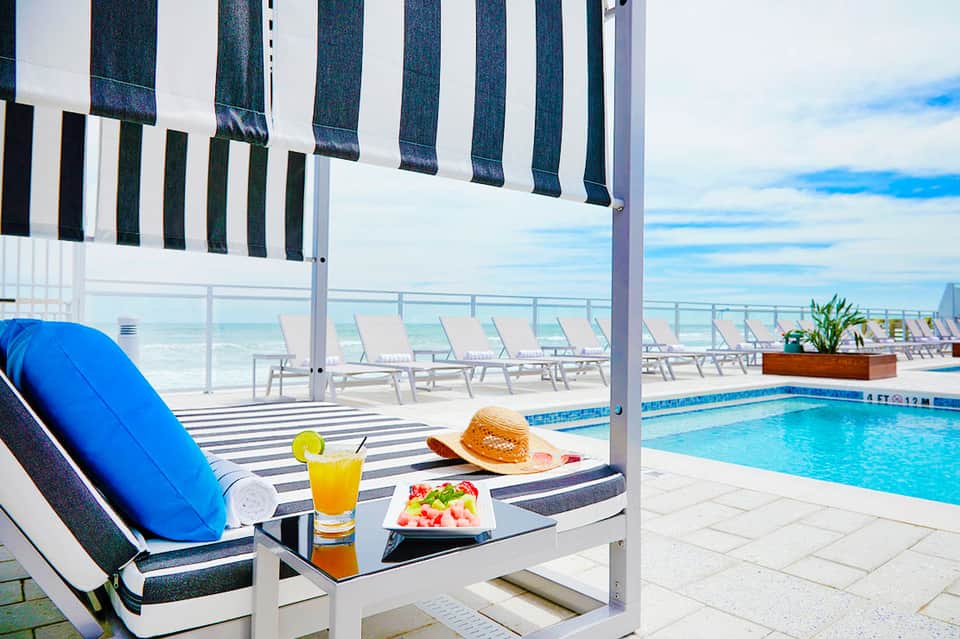 Beachfront cabana with striped awning, lounge chair, refreshments, and ocean view with pool in background