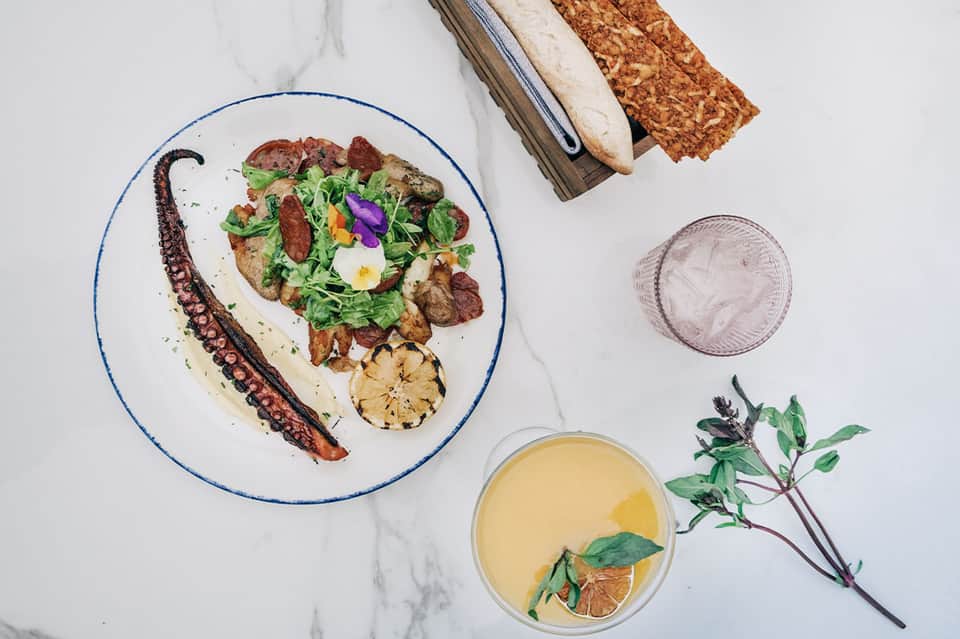 Octopus and salad plated on white marble with bread, juice, and herbs