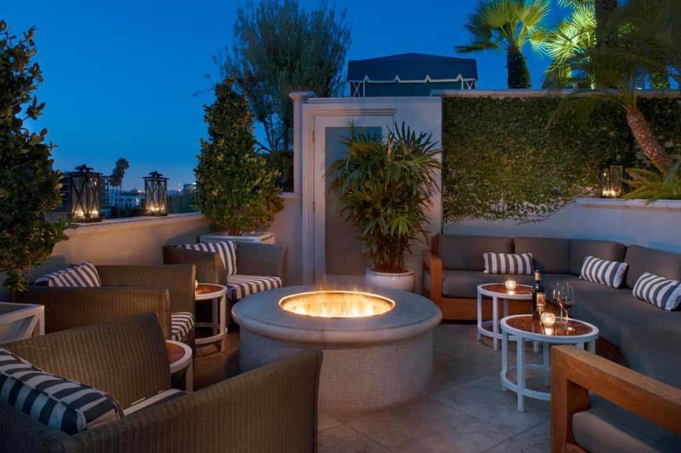 Rooftop lounge at night with fire pit, striped seating, palm trees, and city lights