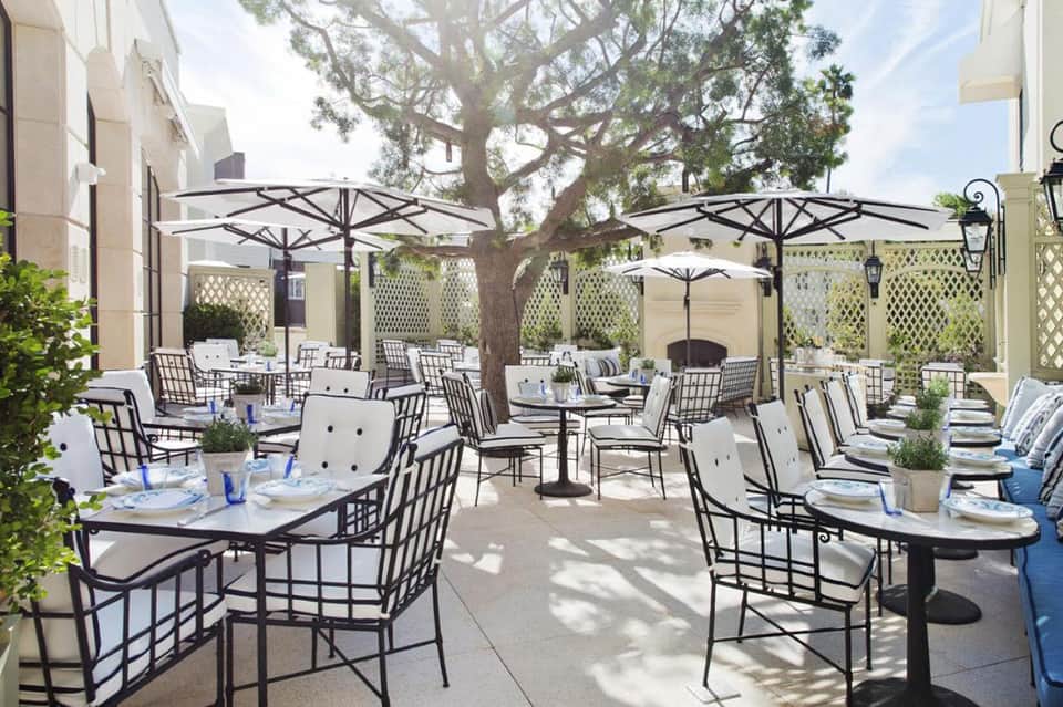Outdoor patio dining area with umbrella-shaded tables, wrought iron chairs, and mature shade tree