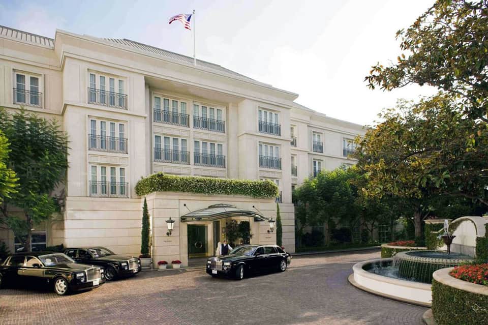 Elegant cream-colored hotel entrance with curved driveway, luxury cars, American flag, and landscaping