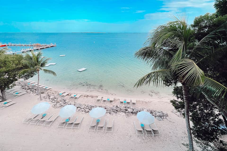 Tropical beach with white sand, palm trees, blue umbrellas and loungers along turquoise ocean
