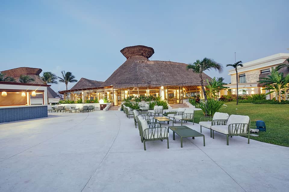 Resort courtyard at dusk with thatched-roof structures, outdoor seating, tropical plants, and modern buildings