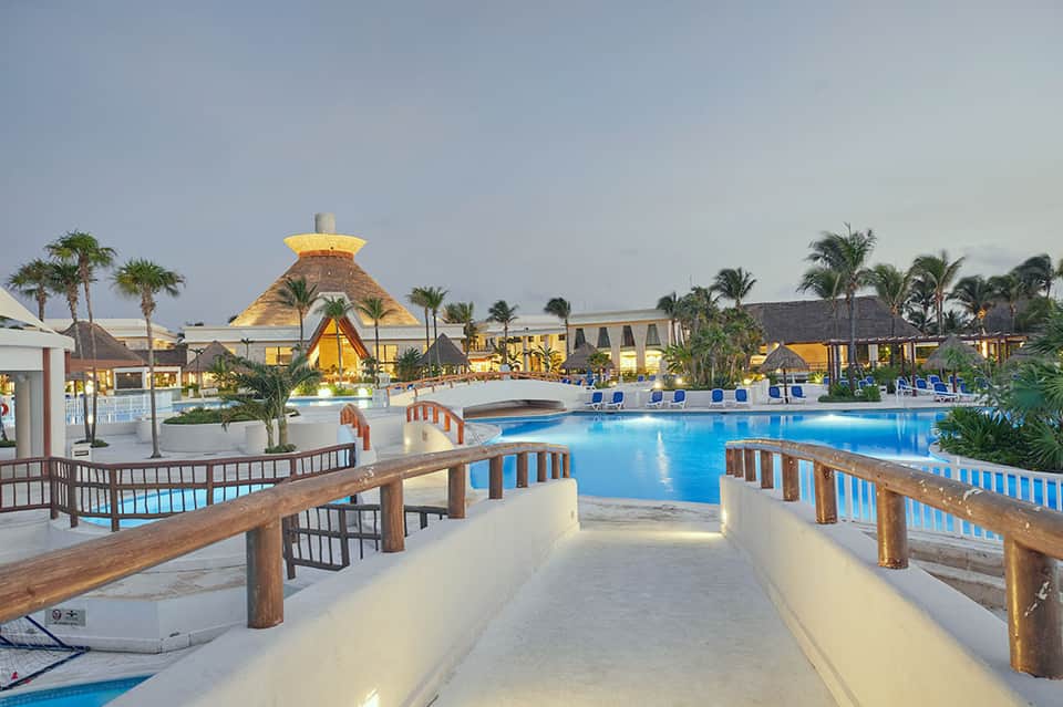 Large resort pool at dusk with wooden bridges, thatched-roof buildings, and illuminated structures