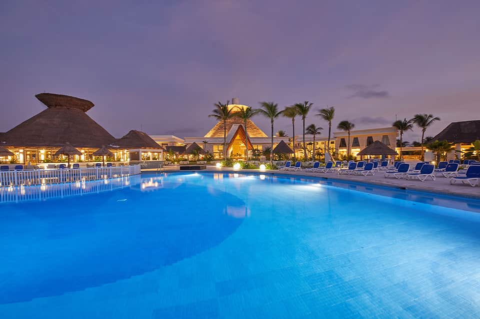 Resort main pool at dusk with illuminated thatched-roof buildings, palm trees, and blue loungers