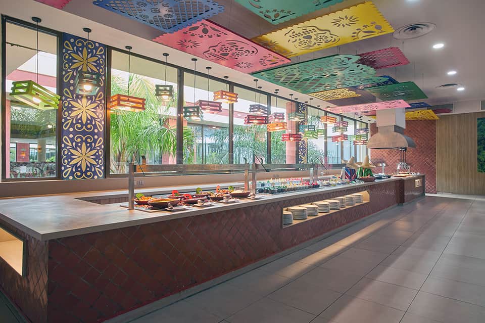 Colorful hotel buffet area with decorative ceiling panels and tropical plants by windows
