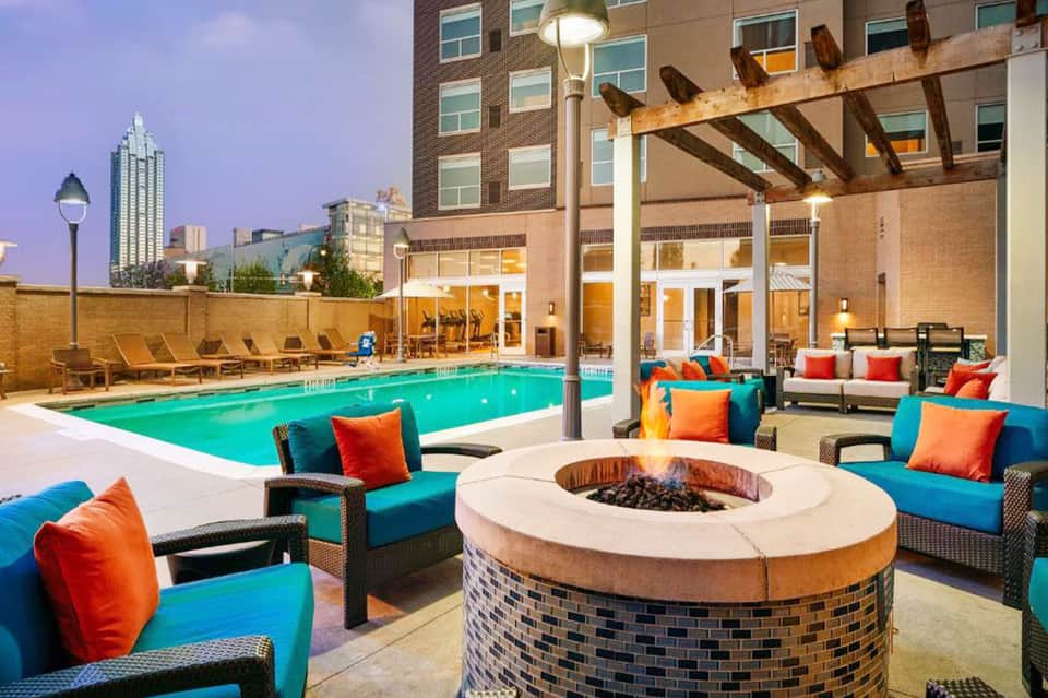 Resort pool deck at dusk with fire pit, lounge seating, turquoise cushions, and city skyline