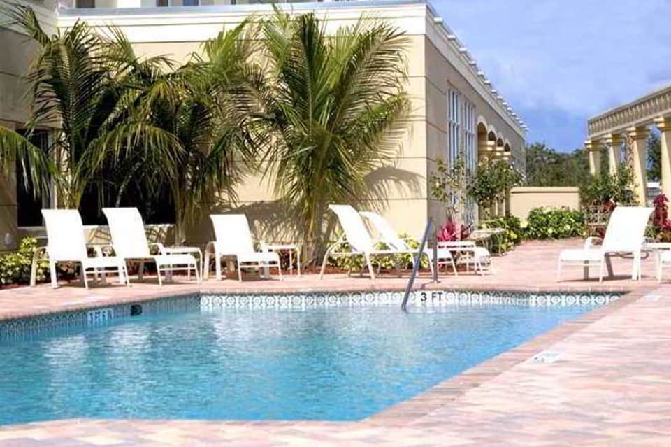 Resort pool with palm trees, white lounge chairs, classical architecture, and tropical landscaping