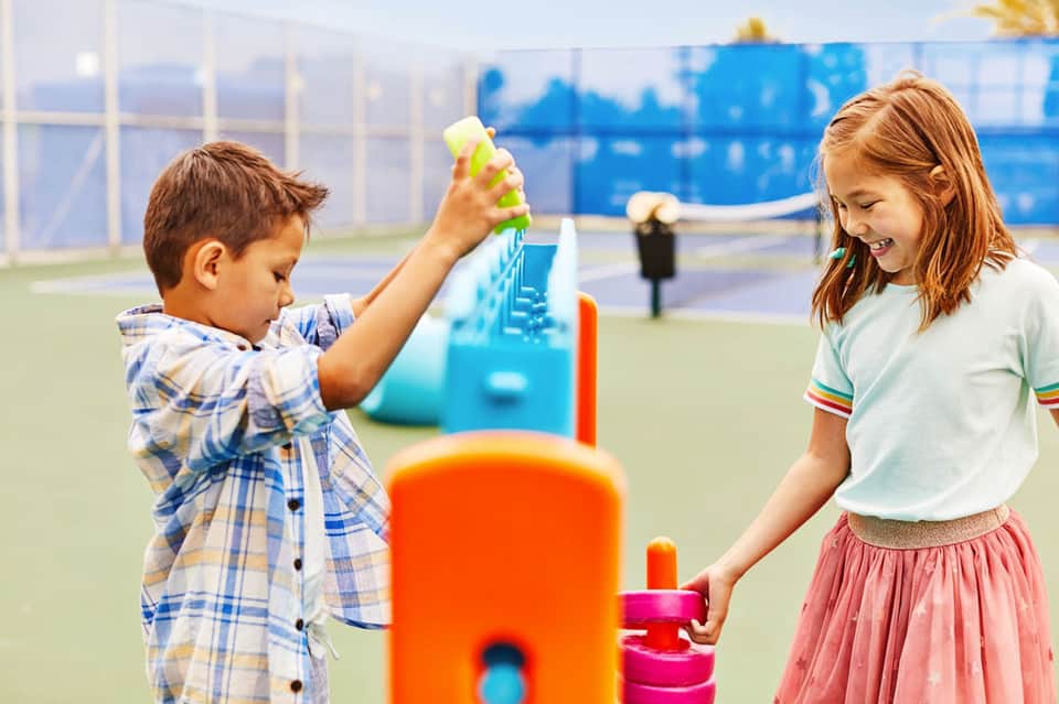 Two children playing with colorful toys on an outdoor tennis court with blue net