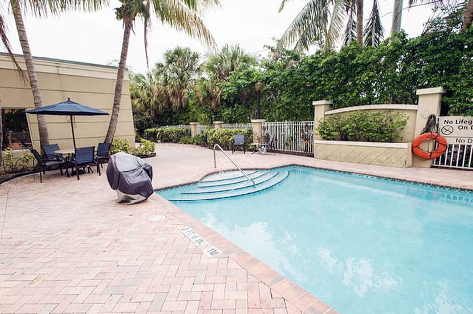 Resort pool with palm trees, lounge chairs, blue umbrella, and enclosed tropical vegetation