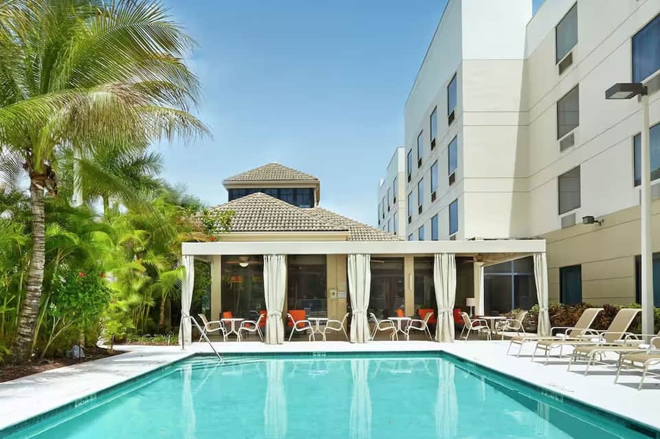 Hotel pool with white pergola cabana, loungers, palm trees, and modern building facade