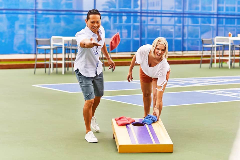 Two people playing cornhole on a tennis court with blue and green courts visible in background