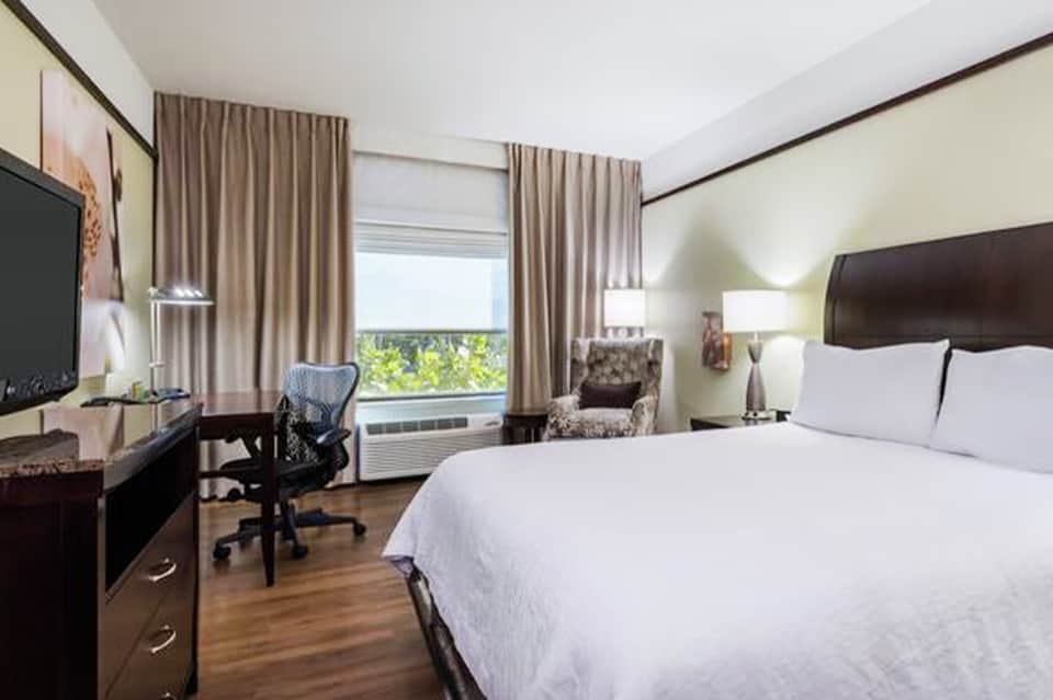 Hotel bedroom with king bed, work desk, and window view of palm trees and waterfront