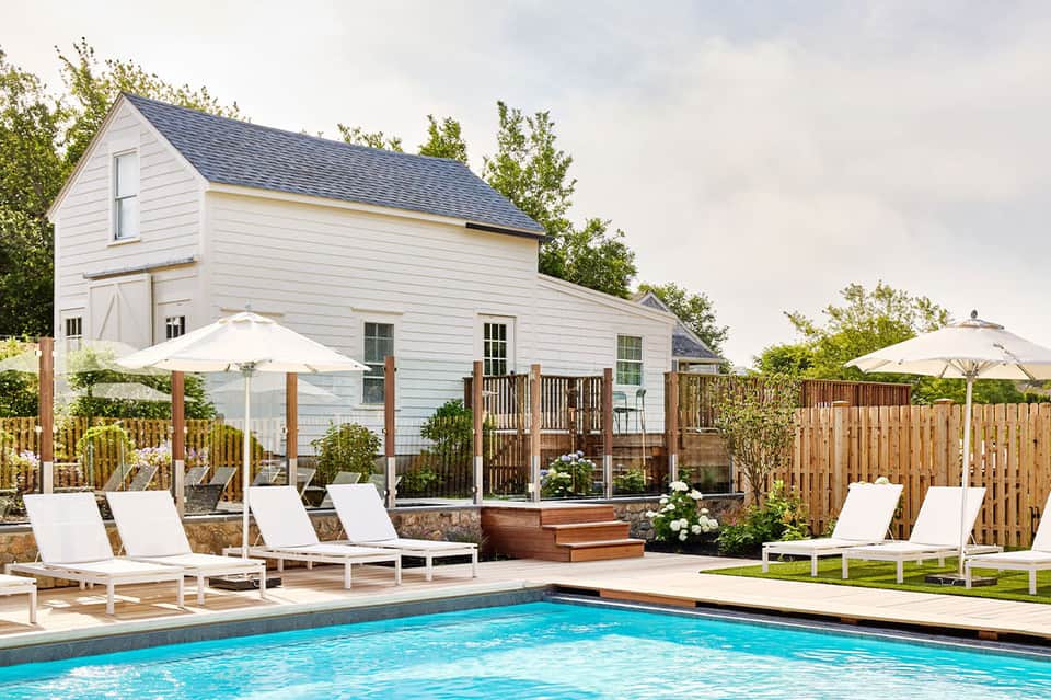 Resort pool with white loungers, umbrellas, and white farmhouse building surrounded by wooden fence