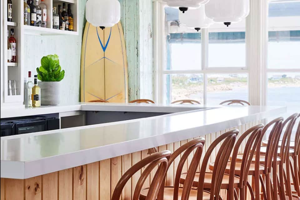 Beachfront bar interior with ocean views, white counter seating, and surfboard wall decoration