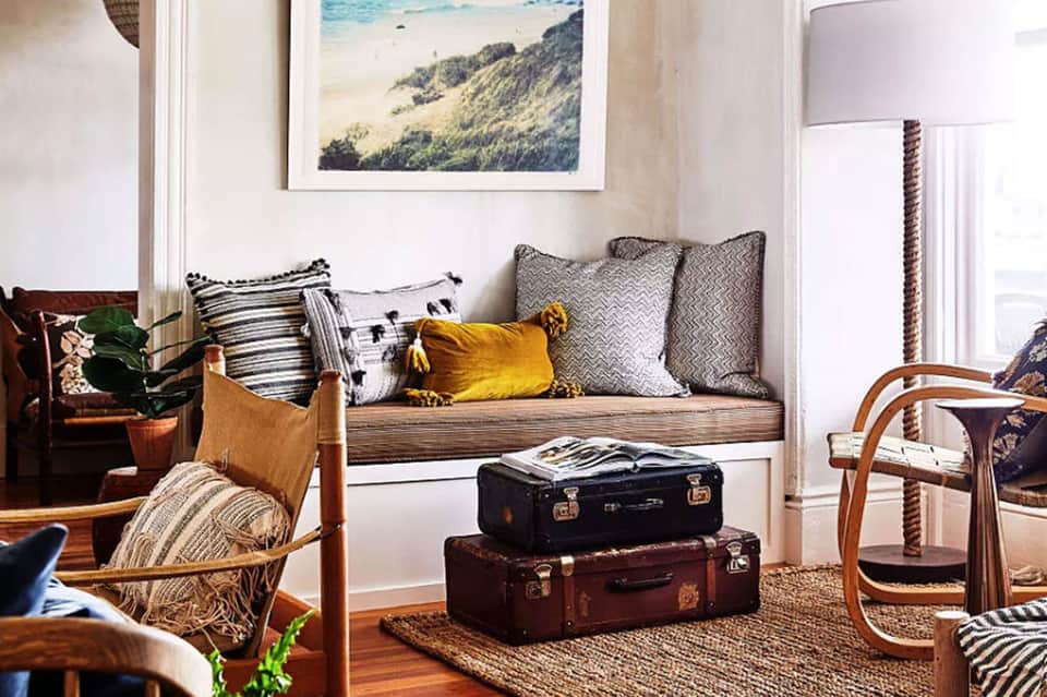 Coastal bedroom with window seat, throw pillows, vintage luggage, and beach landscape art