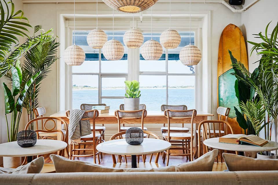 Coastal cafe with woven pendant lights, rattan chairs, white tables, ocean view windows, and tropical plants