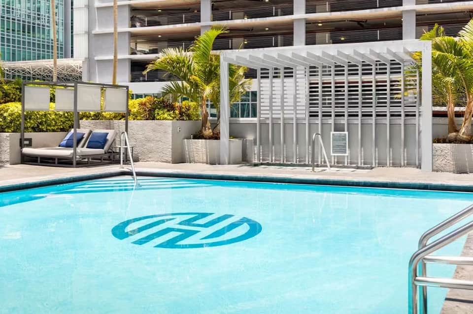 Resort pool with branded logo, modern lounge seating, shaded structures, and landscaping