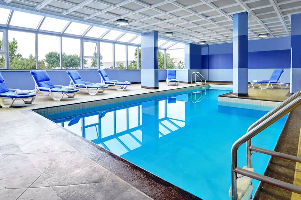 Indoor heated swimming pool with blue loungers and large windows showing natural light