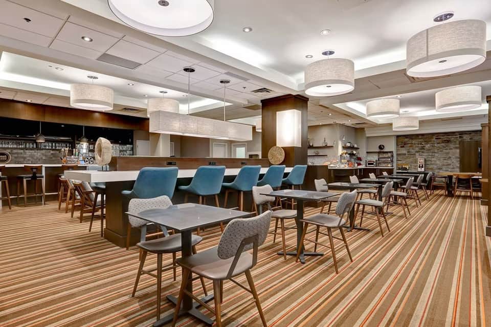 Modern hotel lounge with wood accents, blue chairs, striped flooring, bar area, and pendant lighting