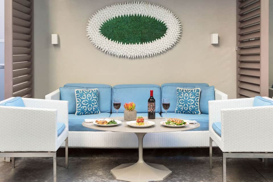Contemporary outdoor lounge with blue sofa, white wicker chairs, artistic green wall art, and dining table