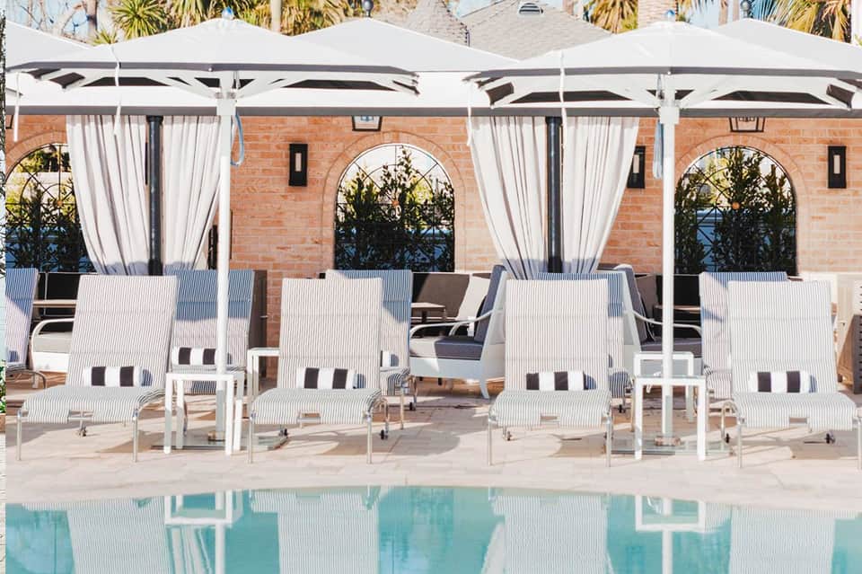 Luxury poolside cabanas with white umbrellas and striped loungers overlooking a turquoise pool against a brick building