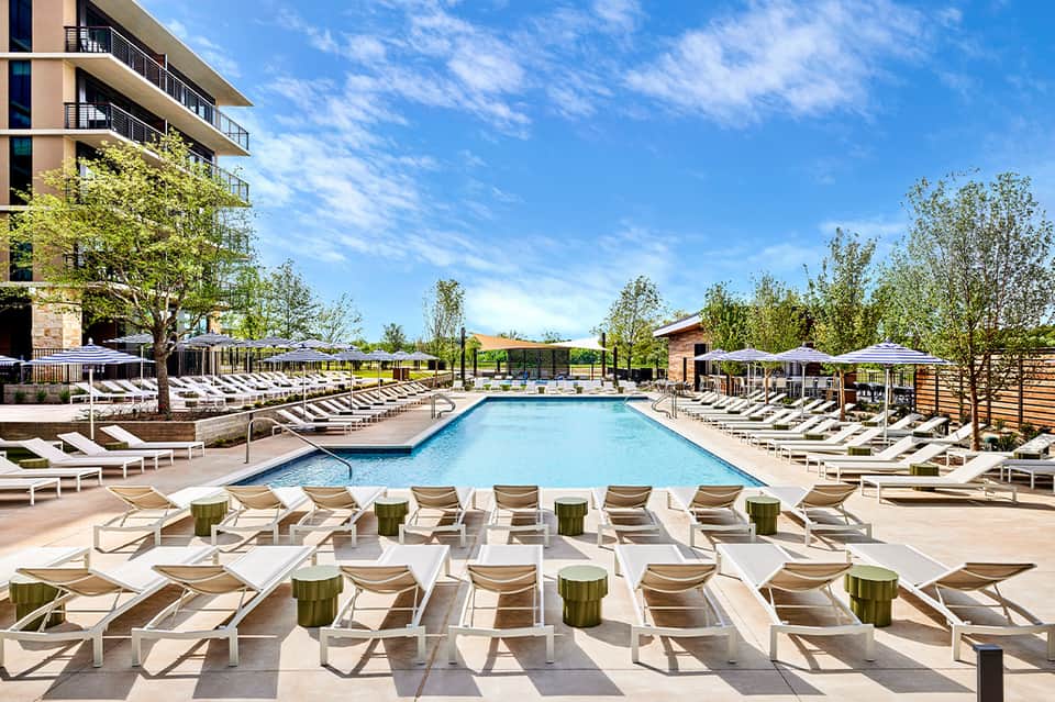 Resort pool surrounded by lounge chairs with umbrellas, modern building, and landscaped lawn areas