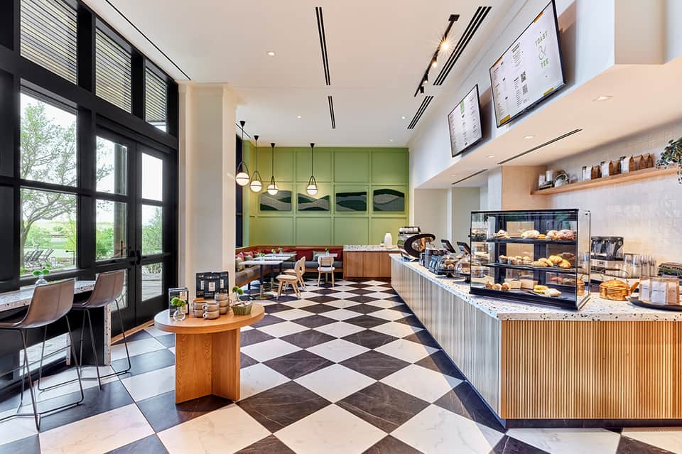 Bright cafe interior with checkered floor, green kitchen, pendant lights, and pastry display