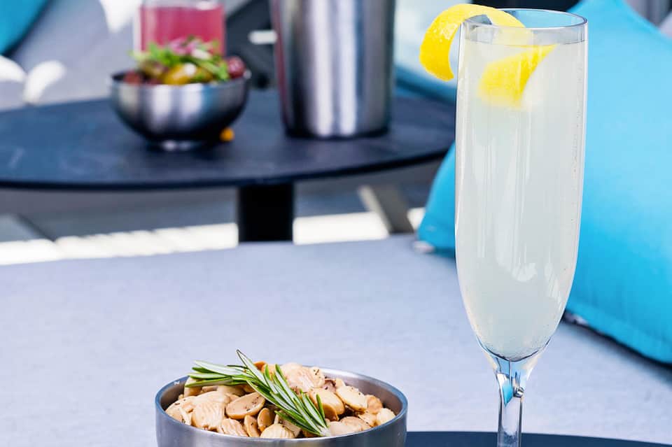 Refreshing lemonade cocktail with lemon twist garnish beside bowl of nuts and appetizers on outdoor table
