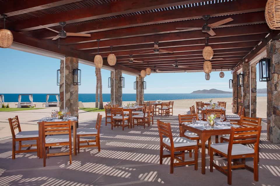 Beachfront restaurant pavilion with wooden tables, stone columns, woven pendant lights, ocean views, and sandy beach access