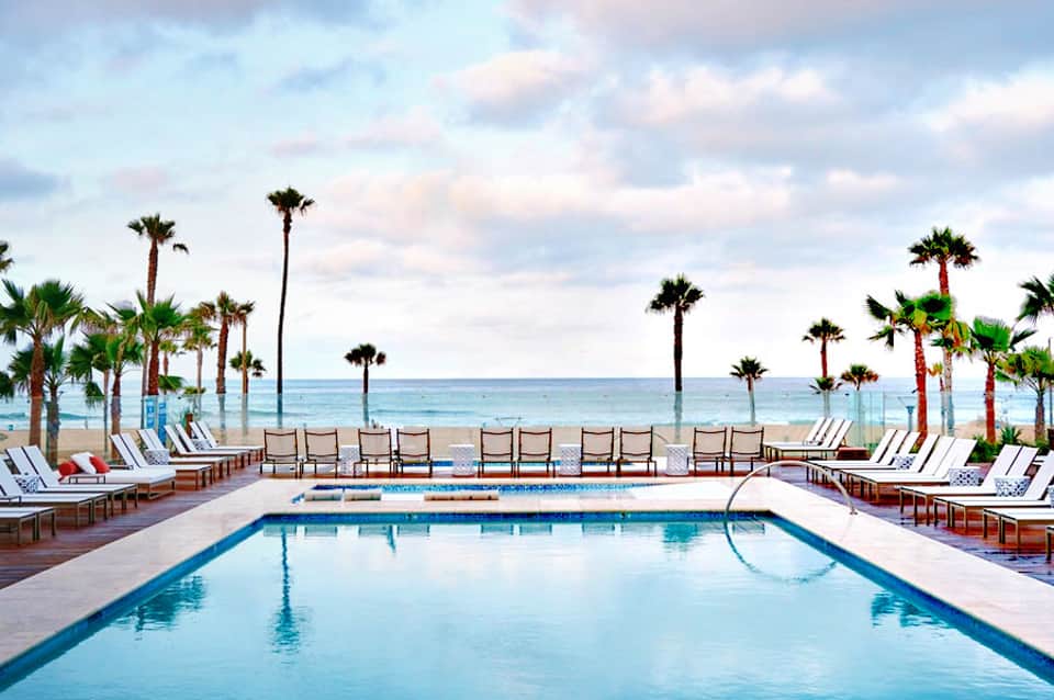 Beachfront resort pool with palm trees, lounge chairs, and ocean view under cloudy sky