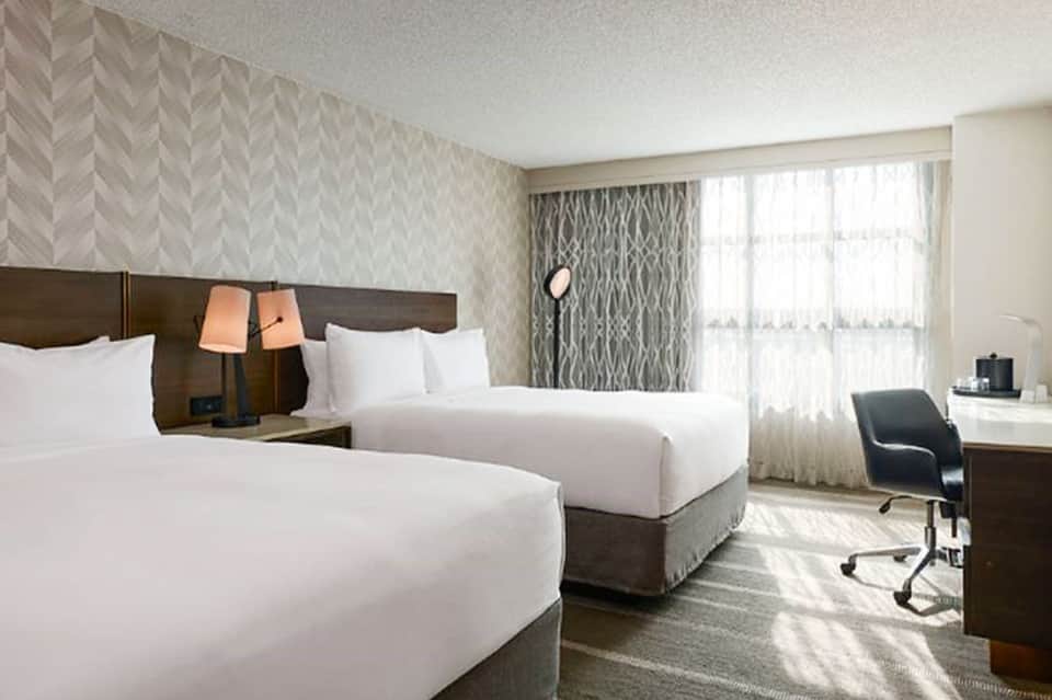 Spacious double queen hotel room with beds, desk, and natural light