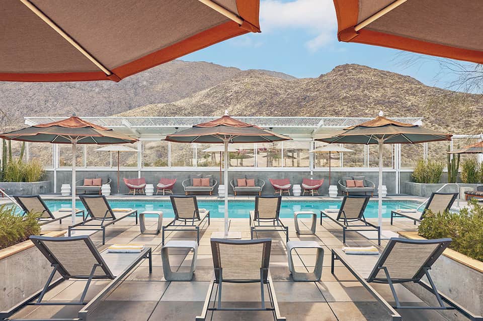 Modern desert resort pool with mountain views, red and tan umbrellas, and contemporary lounge chairs