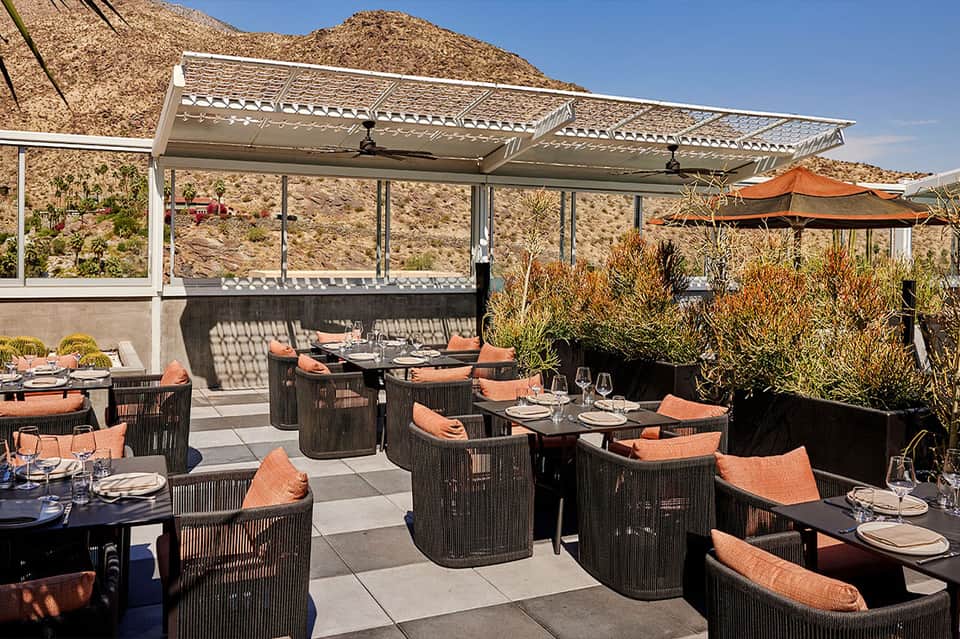 Outdoor dining terrace with dark wicker furniture, orange cushions, and desert mountain backdrop