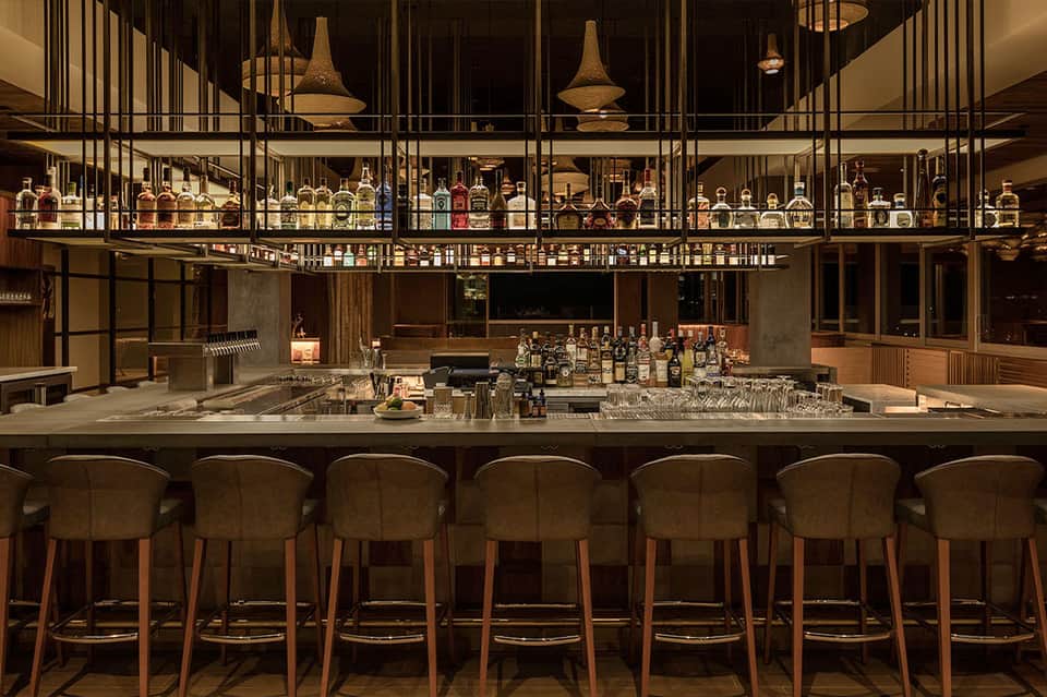 Sophisticated bar with wooden chairs, extensive bottle collection, pendant lights, and modern design