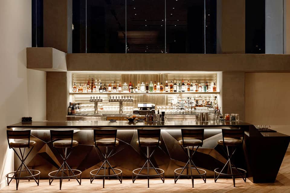 Modern hotel bar with illuminated shelving, bar stools, and floor-to-ceiling windows