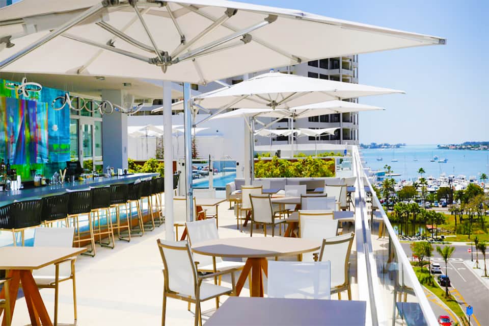 Waterfront restaurant patio with white umbrellas, wooden chairs, bar area, and bay views