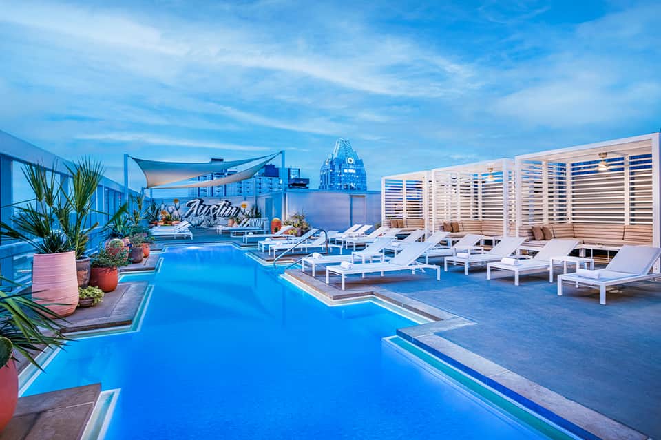 Modern rooftop pool deck with bright blue water, white loungers, privacy screens, and city skyline view