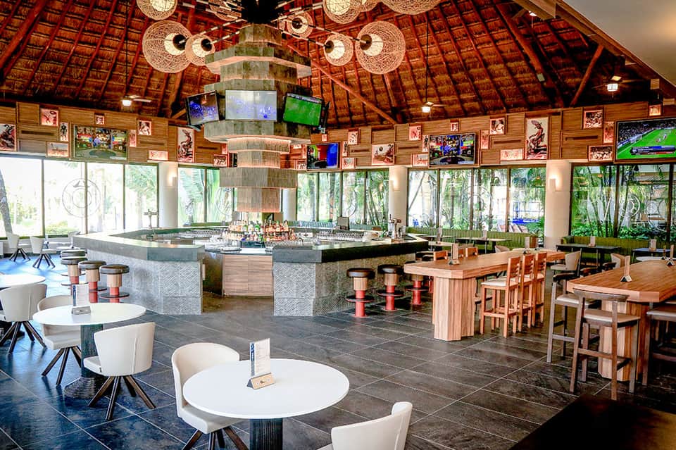 Circular tiki-style bar with rattan ceiling, multiple TV screens, wooden bar seating, and windows