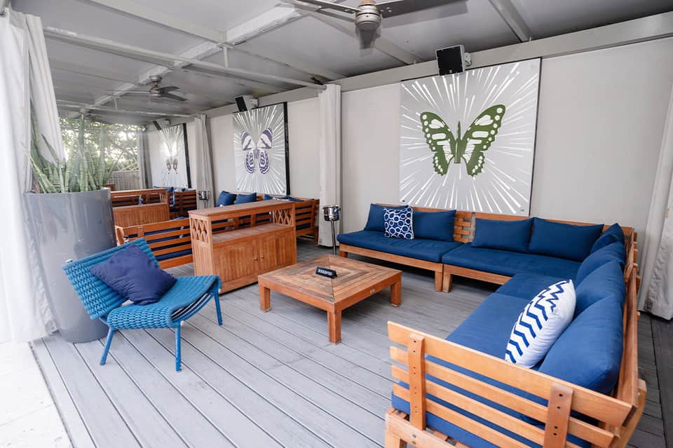 Covered lounge area with wooden furniture, blue cushions, and butterfly artwork on screens