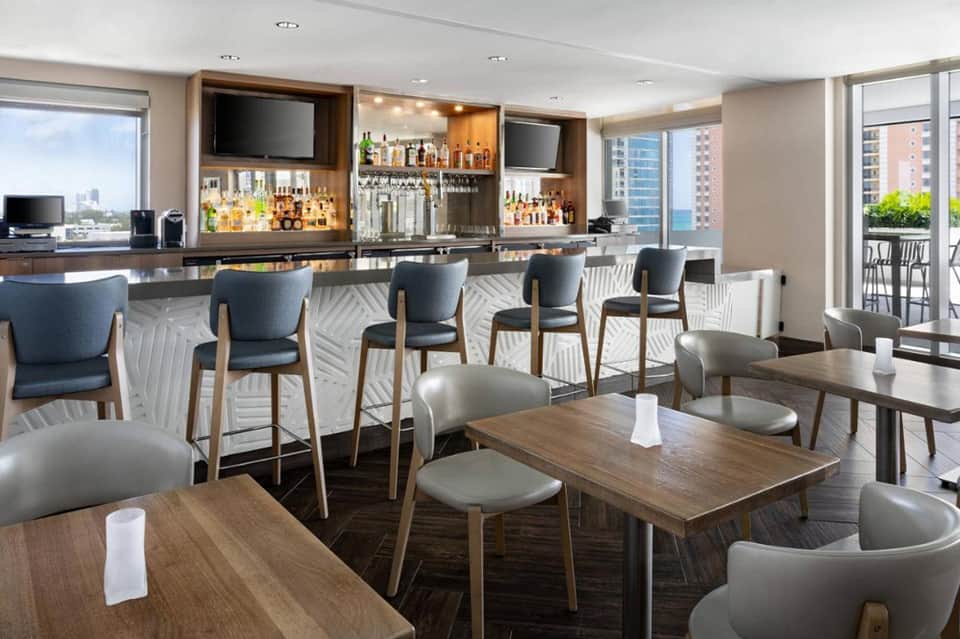 Modern hotel bar lounge with wooden tables, gray chairs, full bar, and floor-to-ceiling windows