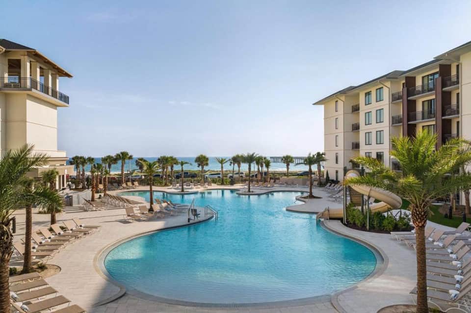 Large curved resort pool with turquoise water, palm trees, and multi-story hotel buildings