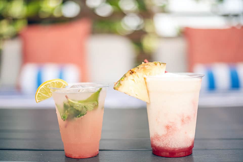 Two tropical cocktails with fresh fruit garnishes displayed on poolside bar table with blurred cabana background.
