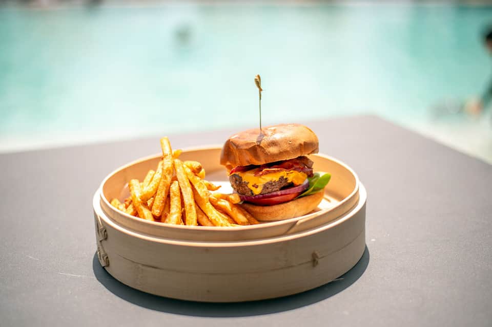 Gourmet burger and fries served poolside on white plate with turquoise pool in background
