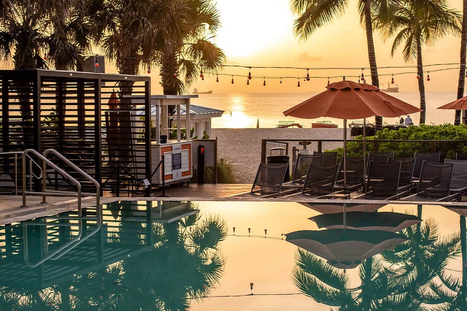 Beachfront resort pool at sunset with umbrellas, lounge chairs, palm trees, and ocean view
