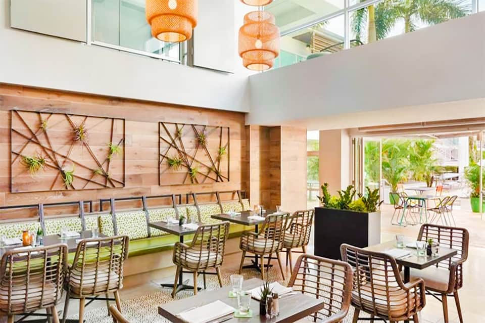Modern restaurant interior with wooden wall art, woven chairs, and orange pendant lights overlooking outdoor seating area