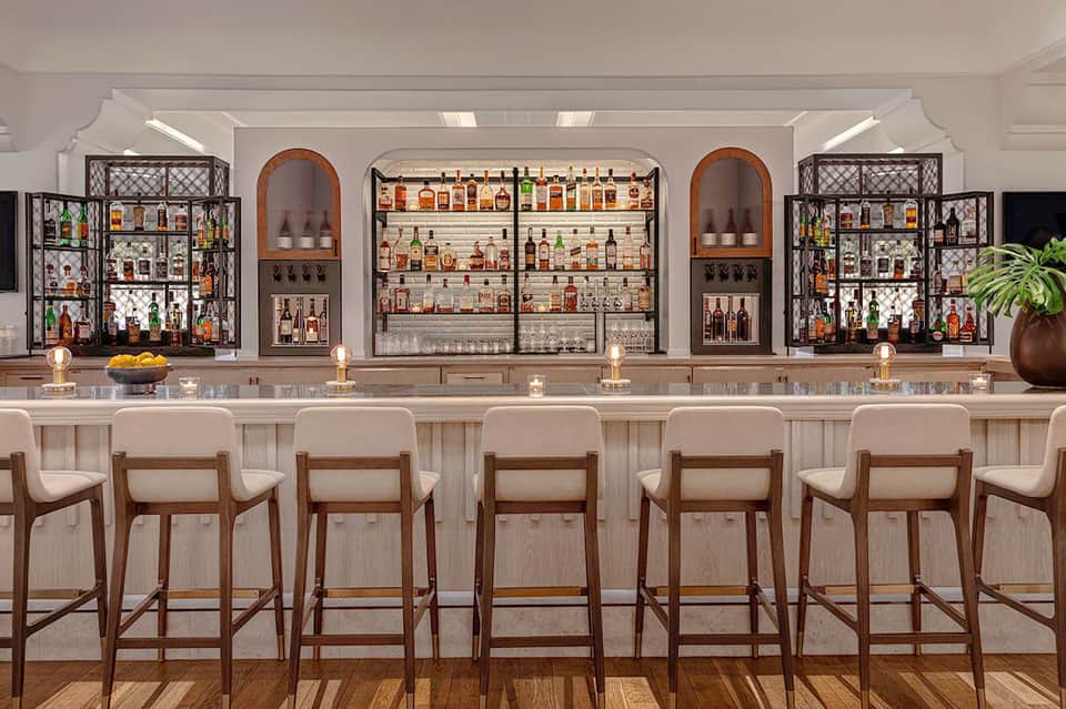 Modern bar with white counter and wood stools, stocked with bottles and decorative shelving