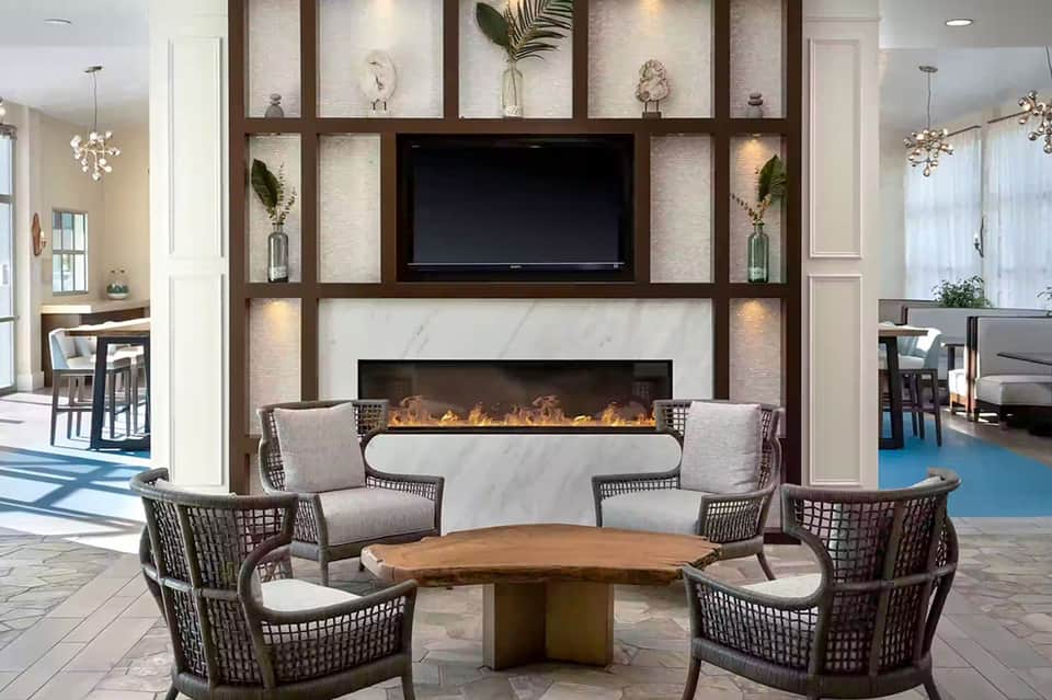 Modern lobby lounge with fireplace, woven chairs, and decorative wall shelving with TV