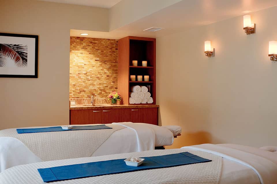 Spa treatment room with massage tables, brick accent wall, wooden shelving with candles and rolled towels
