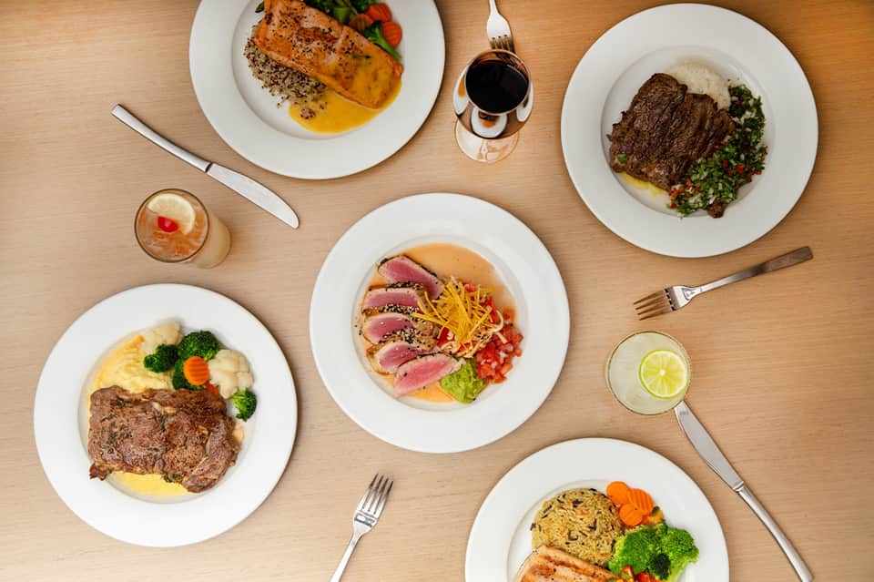 Overhead view of plated fine dining dishes including fish, steak, and seafood with cocktails and wine